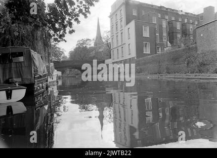 London, England, circa 1967. A view from the Regent’s Canal near Camden, London. Ahead is a building situated in St Mark’s Square and Regent’s Park Road bridge. In the distance is the spire of St. Mark’s Church. Boats are moored on the canal and a couple are walking along the towpath. Stock Photo