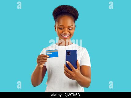 Young happy African American woman uses credit card to shop on mobile phone, stands in studio Stock Photo