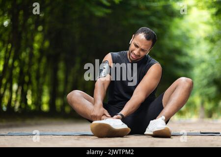Handsome black sportsman exercising in public park, touching ankle Stock Photo