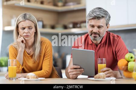 Suspicious lady watching her husband using digital tablet Stock Photo