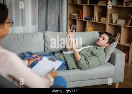 Sad young european guy patient lies on sofa and talks with black woman doctor in office
