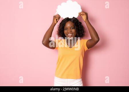 Close up photo of funny pretty dark skin lady holding paper cloud mind thinking over creative dialogue answer doubtful wear orange shirt isolated pink Stock Photo