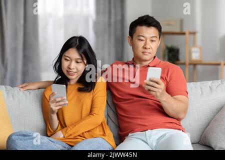 Asian couple sitting on couch together, using their smartphones Stock Photo