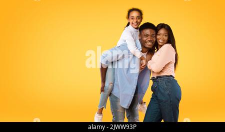 Happy young african american family posing, man giving piggy back ride for his daughter, yellow background, free space Stock Photo