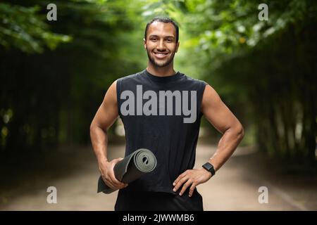 Joyful black guy having workout at public park, holding mat Stock Photo