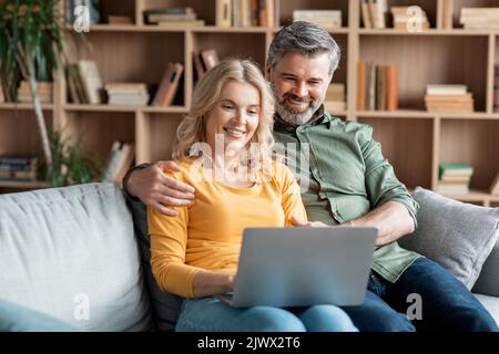Smiling Middle Aged Spouses Resting With Laptop In Living Room Interior Stock Photo