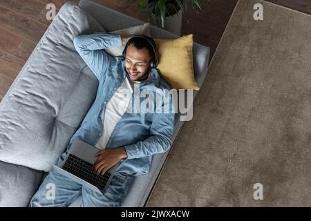 Young Black Man Wearing Headphones Relaxing On Couch With Laptop, Top View Stock Photo