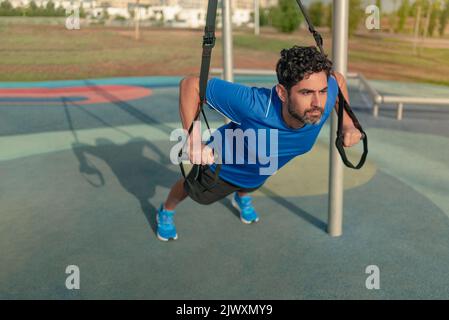 Young man doing fitness exercises on a special hanging device. Fitness male guy doing exercising with suspension trainer slingin a city park. A suspen Stock Photo