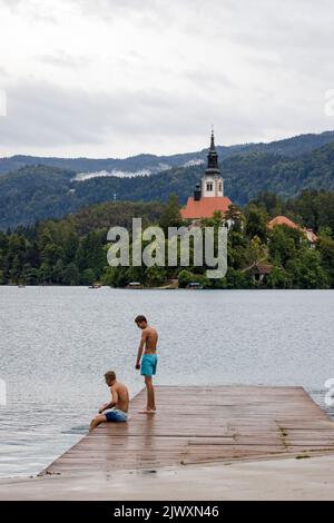 Lake Bled in Slovenia with the church and two boys ready to go swimming from the pier in the foreground Stock Photo