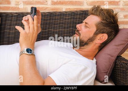 Relaxed casual man using a smart phone lying on the couch at the terrace. Stock Photo