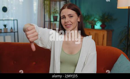 Dislike. Upset girl showing thumb down sign gesture, expressing discontent, disapproval, dissatisfied bad work at modern home apartment indoors. Displeased young woman in living room sitting on sofa Stock Photo