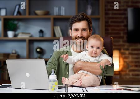 Portrait of a Father on maternity leave, in his home office with a small child in his arms. Dad at the table with a laptop and a child. Holiday to Stock Photo