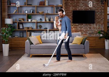 A young father, with a child, does housework. Father on maternity leave. Dad cheerfully sings, dances and vacuums, cleans with a small child in his Stock Photo