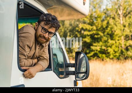 Cheerful young bearded man leaning out of the window of a camper van and smiling ar the camera. . High quality photo Stock Photo