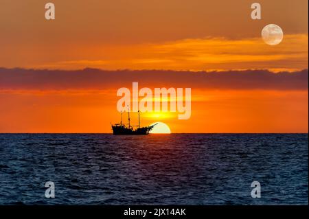 A Silhouette Of A Pirate Ship Sits At Sea As The Sun Rises On the Ocean Horizon Stock Photo