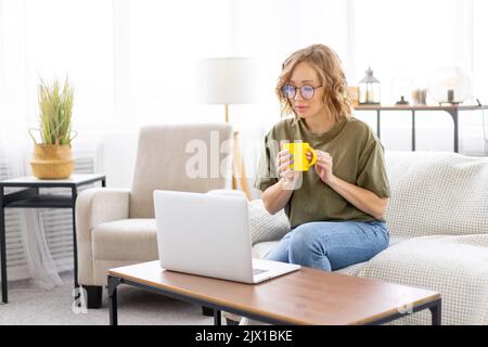 Woman with glasses use laptop typing keyboard sitting couch big window background home interior Freelance female working from home Distance learning student relaxing watch lessons video conference. Stock Photo