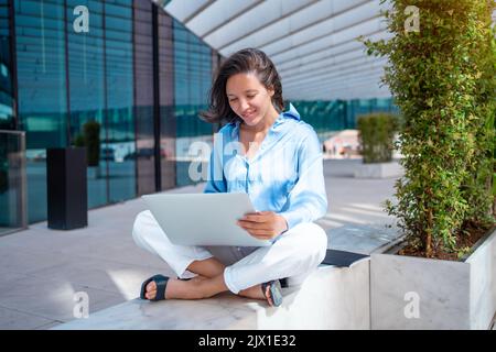 Pretty business woman use laptop outside sitting lotus position near office building. Positive smile caucasian female business person do remote work outdoors. Self employed freelance worker Stock Photo