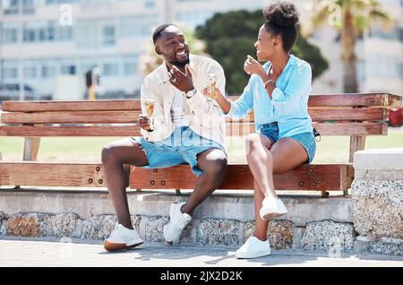 Happy black couple eating ice cream on a beach bench together, smiling while bonding and laughing. Young African American man and woman enjoying their Stock Photo