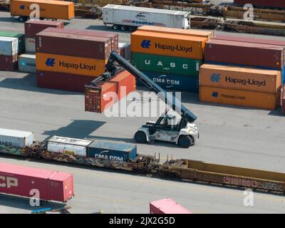 An aerial photo of a shipping container being moved by a large industrial forklift in a shipping terminal. Stock Photo
