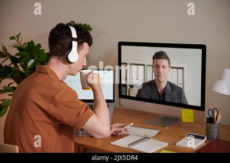Young man drinking coffee when talking to coworker at online meeting Stock Photo