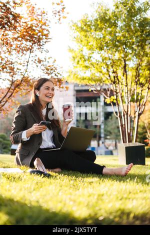 Beautiful woman sitting on grass lawn at park holding a coffee and a smartphone, searching information for her work on laptop. Business success Stock Photo