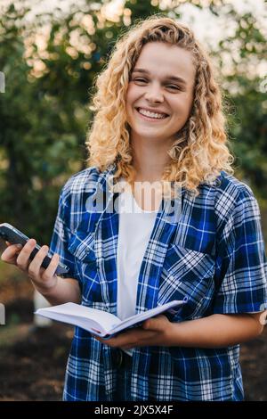 Smiling caucasian curly-haired woman holding mobile phone and notebook to collect data about agriculture problems. The concept of agricultural farms Stock Photo