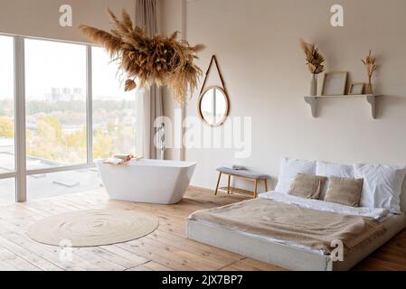 Bright modern boho chic bedroom with a freestanding white bathtub against a panoramic window and dry grass and flowers hanging decor. Soft selective f Stock Photo