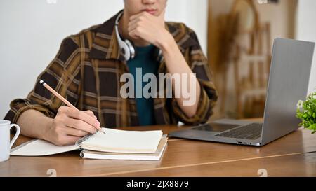 Thoughtful young Asian male freelancer or college student working at his desk, thinking pensively to plan his project, doing homework. cropped image Stock Photo