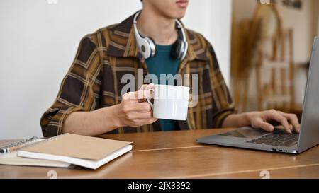 A handsome young Asian male college student or freelancer with wireless headphones, working on his project on laptop while sipping a morning coffee. c Stock Photo