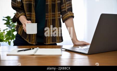 Professional young Asian male freelancer or programmer in flannel shirt leaning on his office desk, holding a mug of coffee. cropped image Stock Photo