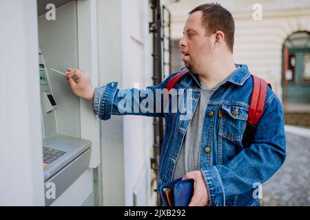 Happy young man with Down sydrome using a street ATM machine and withdrawing money. Stock Photo