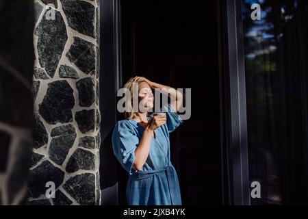 Happy young woman standing in garden near cottege and enjoying cup of morning coffee on summer vacation in mountains. Stock Photo