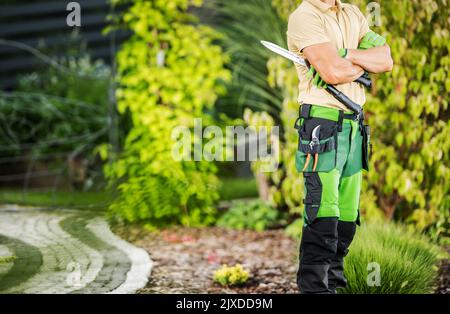 Thoughtful Caucasian Gardener with Folded Hands Standing in the Garden Getting Ready to Start Trimming and Shaping Work Using Pruning and Hedge Shears Stock Photo