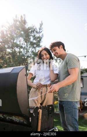 Smiling man cooking on grill near girlfriend with wine outdoors,stock image Stock Photo