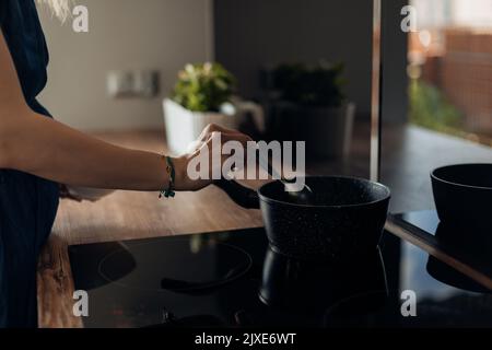 Close up of cropped unrecognizable woman hands cooking food in frying pan on induction cooker, stove with ceramic top Stock Photo