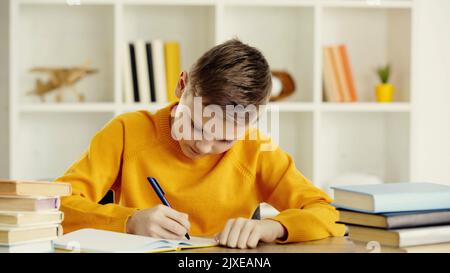 preteen boy writing in notebook while doing homework near books on wooden table at home,stock image Stock Photo