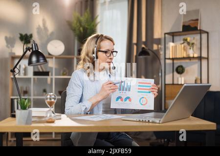 Smiling caucasian woman showing graphs and charts on laptop computer during video conference sitting at bright office or home. Pretty female software developer using modern laptop for remote work. Stock Photo