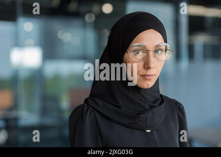 Close-up photo of business woman in black hijab, muslim woman working at work inside modern office building, looking at camera with concentration and serious thoughtfulness in glasses. Stock Photo
