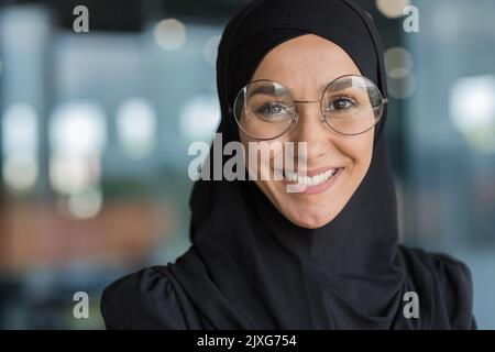 Close-up portrait of Muslim businesswoman in hijab smiling and looking at camera, female worker in glasses working inside modern office building. Stock Photo