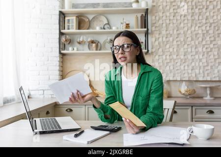 Mortgage loan. Upset and angry young woman holding a rent bill in her hands. Received a letter about the debt. Sittin g in the kitchen at home at the table with a laptop in glasses and a green jacket Stock Photo