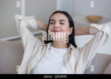 Close up photo. Happy young beautiful woman relaxing at home on sofa with eyes closed, arms behind head. Enjoys silence, peace, dreams, smiles. Have a nice weekend. Stock Photo