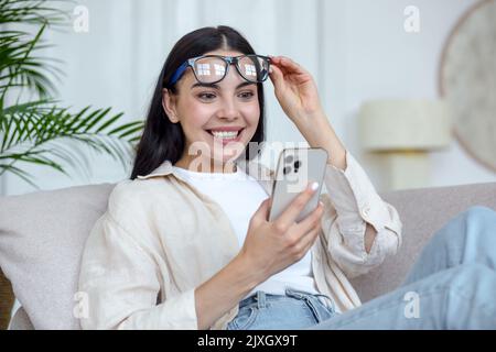 Happy young woman in glasses sitting on sofa at home, holding mobile phone, reading message, got good news. Satisfied and surprised, she lifted her glasses from her face with her hand. He smiles. Stock Photo