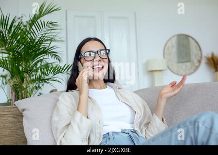 Happy young beautiful woman in glasses sitting on the sofa at home and talking on the phone with friends and family. Gesturing with hands. Quarantine, remote online communication. Stock Photo