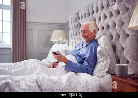 Smiling Senior Man At Home Wearing Pyjamas In Bed Using Mobile Phone Stock Photo