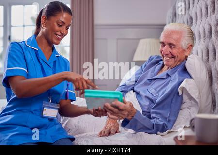 Female Care Worker In Uniform Helping Senior Man At Home In Bed With Medication Stock Photo