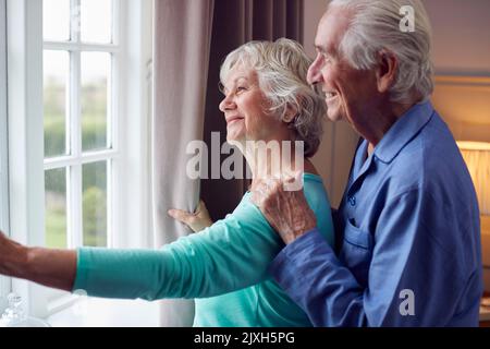 Smiling Senior Couple At Home Wearing Pyjamas Opening Bedroom Curtains And Looking Out Of Window Stock Photo