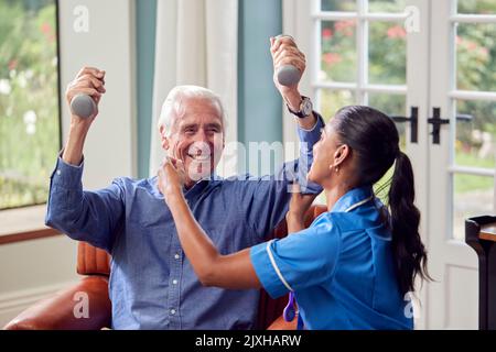 Female Nurse Or Physiotherapist In Uniform Helping Senior Man To Lift Hand Weights At Home Stock Photo