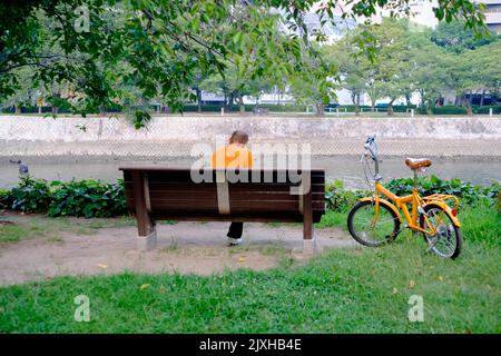 Elderly person enjoys sitting on the park bench after riding his orange bicycle. Stock Photo