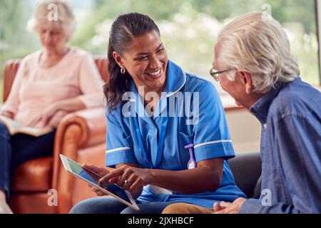 Senior Couple At Home With Man Talking To Female Nurse Or Care Worker Using Digital Tablet Stock Photo