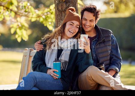 Smiling Couple Sitting On Bench In Autumn Park Using Mobile Phone Stock Photo